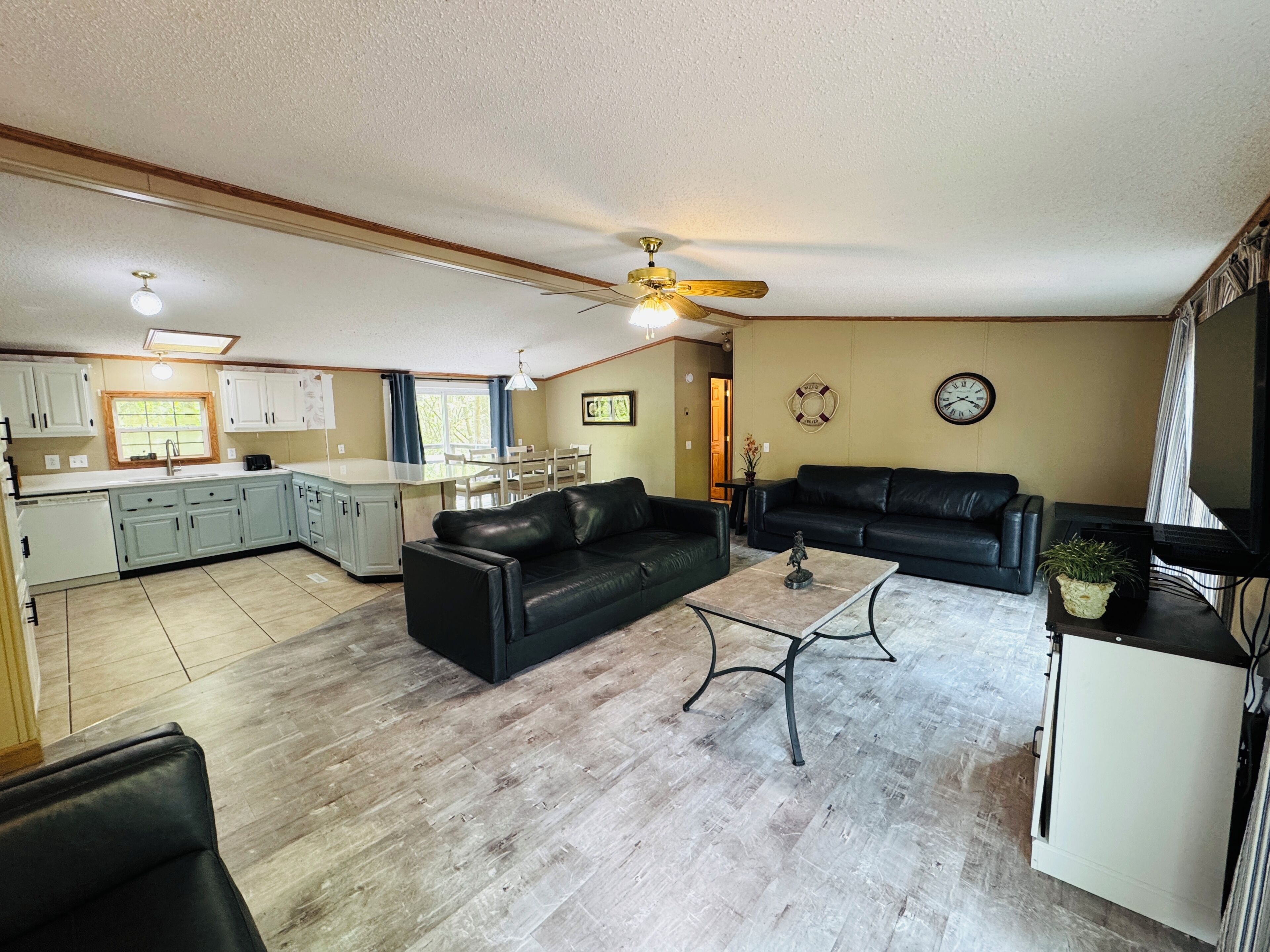 A living room with a black leather sofa, a coffee table, and a TV. There is a kitchen area with white cabinets and a dining table.