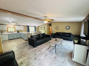 A living room with a black leather sofa, a coffee table, and a TV. There is a kitchen area with white cabinets and a dining table.