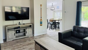 A living room with a TV, wooden entertainment unit, black leather sofa, and a dining area with a table and chairs. - Chill & Cozy Back Escape—Grill Time | 8 ppl | IC99 (Put-in-Bay)