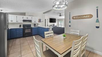 A modern kitchen with white and blue cabinets, a wooden dining table with chairs, and a 'WELCOME' sign on the wall.