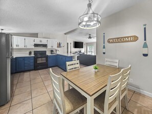A modern kitchen with white and blue cabinets, a wooden dining table with chairs, and a 'WELCOME' sign on the wall.