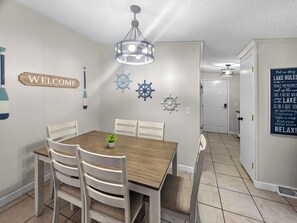 Un comedor con una mesa de madera y sillas blancas, un cartel de bienvenida, decoración de pared con temática náutica y un pasillo con un letrero que dice 'Lake Rules'.