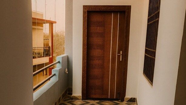 A narrow hallway with a patterned floor, a wooden door, and a window overlooking a balcony. - SRT Homes (Ranchi)