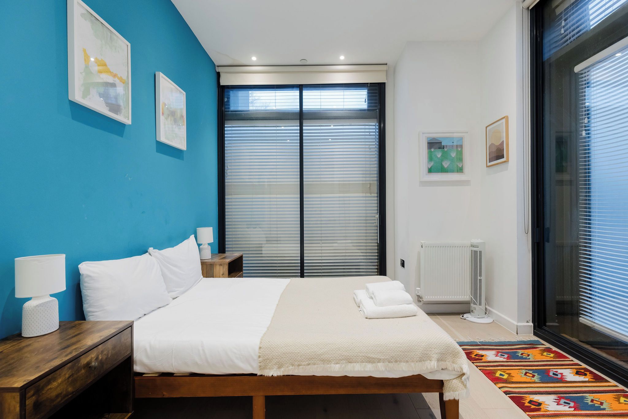 A bedroom with a bed, bedside tables, a radiator, and a window with blinds.