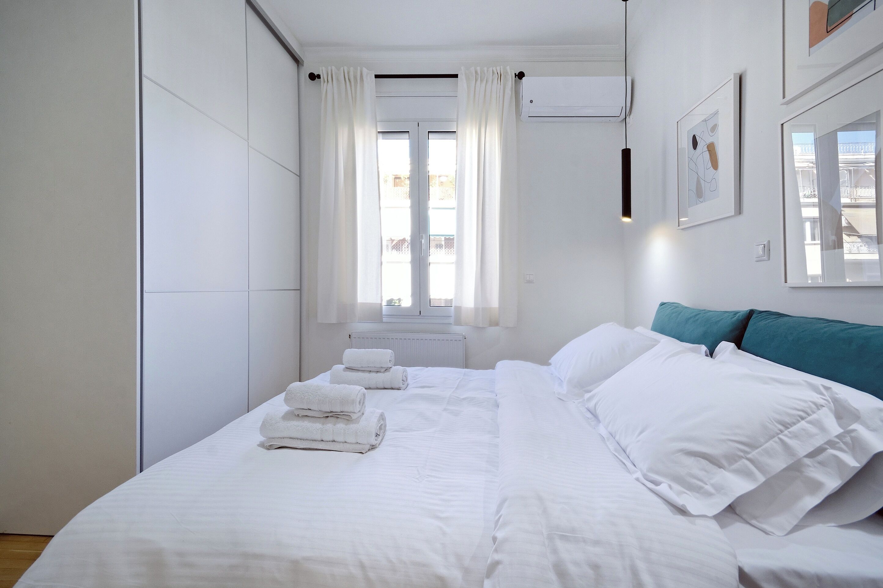Un lit bien fait, avec du linge de lit blanc et deux serviettes enroulées, une fenêtre avec des rideaux blancs et un coussin turquoise sur le lit.