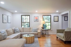A living room with a sectional sofa, a round coffee table, and two armchairs. There are two windows with views of trees, a lamp on a side table, and framed artwork on the walls. - Dog Friendly, Game Room, Central AC! (Chatham)