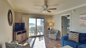 A living room with a TV, a ceiling fan, a sofa, a patterned armchair, and a view of the ocean through a sliding glass door. - Regency Towers #407 East | Pet Friendly, Beach Service, Pool, Pickleball (Pensacola Beach)