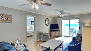 A living room with a ceiling fan, a TV, and a view of the ocean through a sliding glass door. - Regency Towers #407 East | Pet Friendly, Beach Service, Pool, Pickleball (Pensacola Beach)