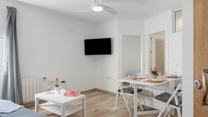 A modern, well-lit room with a flat-screen TV mounted on the wall, a dining table with chairs, a small white table with a vase and a book, and a bed with a gray blanket. - Belvedere Apartment with Views of Vejer’s Historic Old Town (Vejer de la Frontera)
