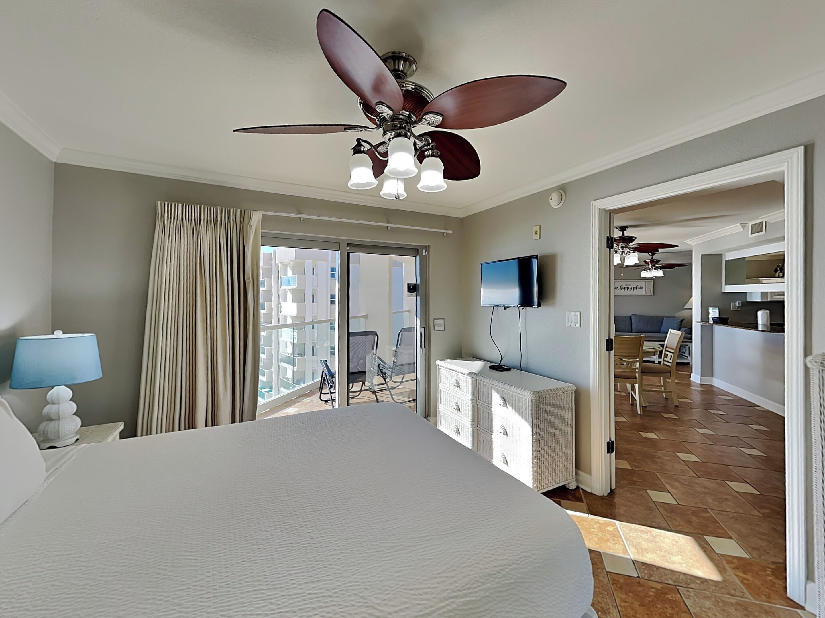 Habitación de hotel con una cama grande, ventilador de techo, televisor, cómoda y vistas al balcón y edificios exteriores.