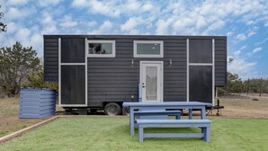Una pequeña casa móvil de color oscuro con una puerta principal, dos ventanas y una mesa de picnic azul en la parte delantera.