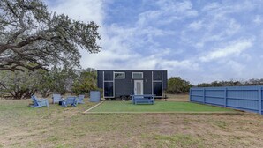 Una casa pequeña de color oscuro con puerta principal y ventanas, rodeada por un jardín césped y una cerca azul.