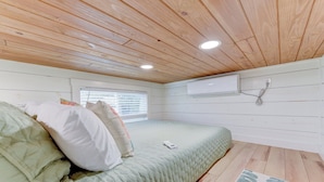 Quarto com cama, almofadas, janela com persianas, aparelho de ar condicionado e teto de madeira.