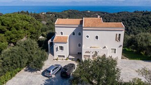 A large, two-story building with a tiled roof, surrounded by greenery and a view of the sea.