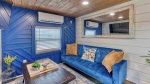 Ein Zimmer mit blauer Wand, einer Holzbodenverkleidung, einem blauen Sofa mit Kissen, einem Holztischchen, einem Spiegel und einer Klimaanlage.