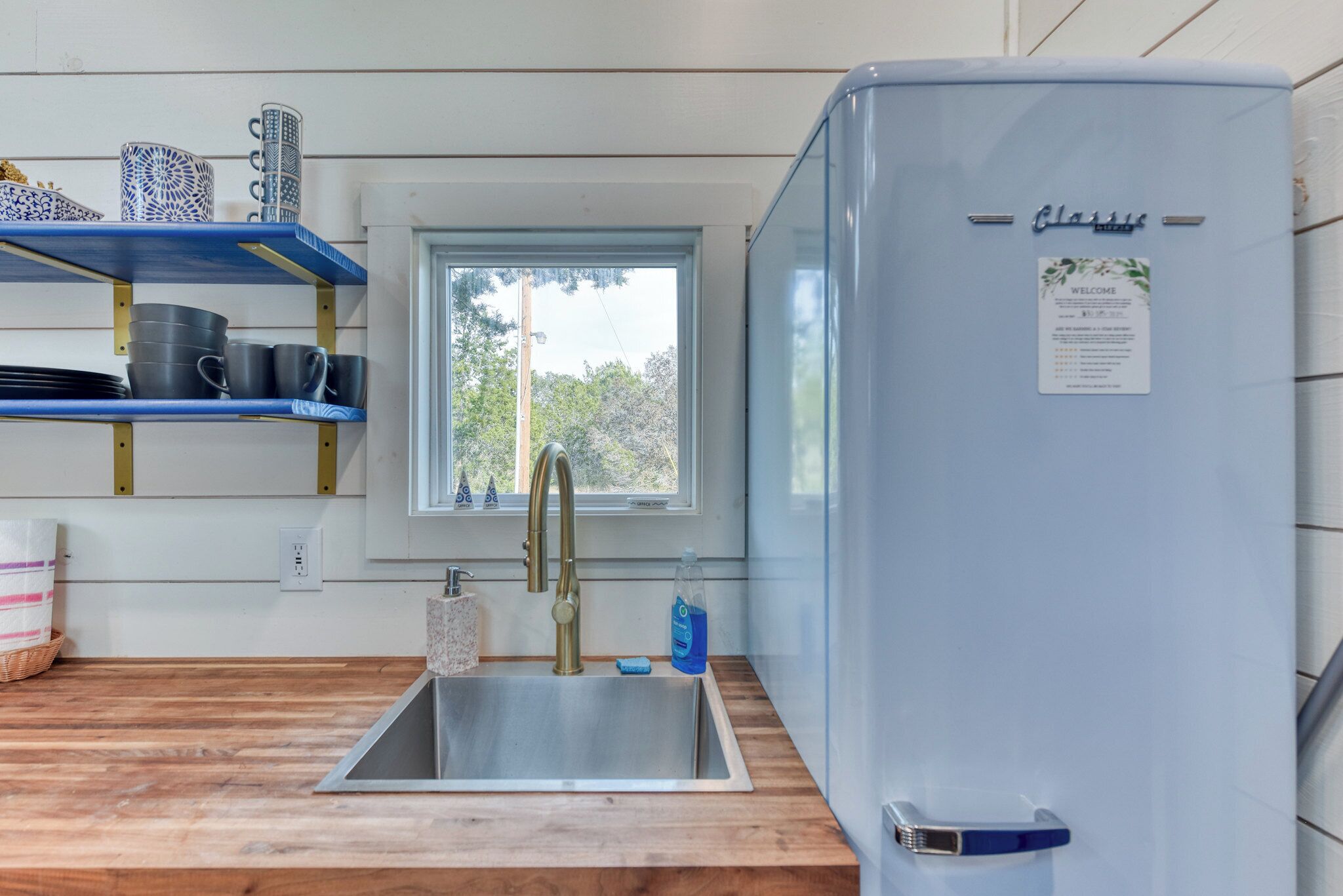 Una cocina con un lavamanos de acero inoxidable, un refrigerador azul y estantes con platos y tazas.