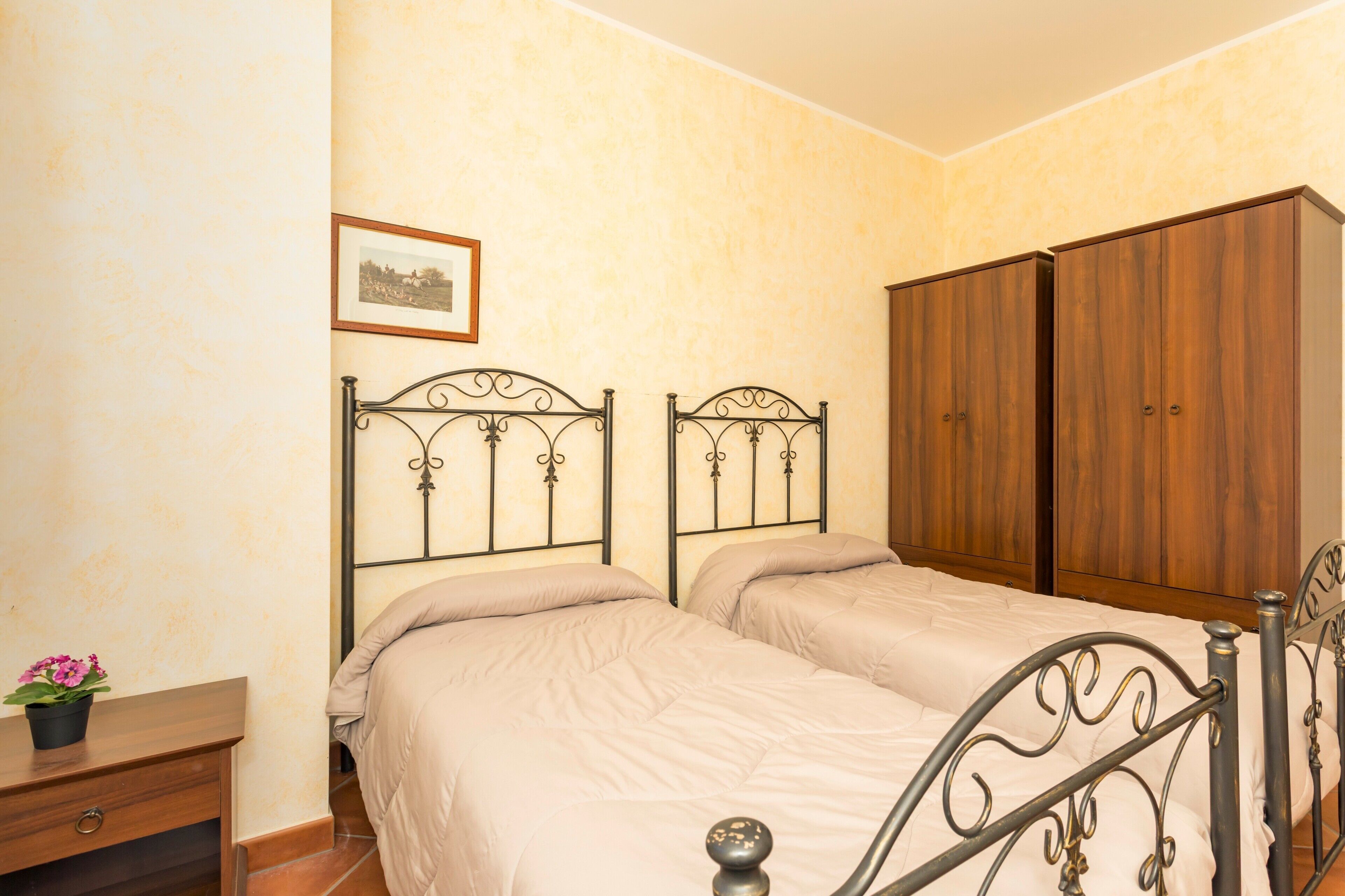 Une chambre avec deux lits, une armoire en bois, une table de chevet avec une plante et un tableau encadré au mur.