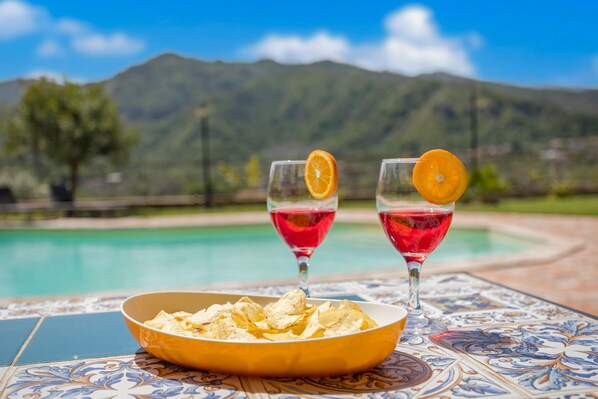 A poolside table with a bowl of chips and two glasses of red drink, with a mountain view in the background.