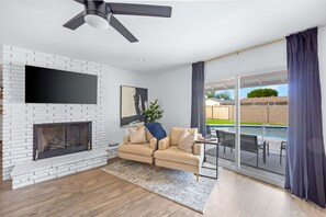 A modern living room with a fireplace, a flat-screen TV, a sofa, and a view of a swimming pool through large glass doors. - Single-Level Home | Pool, Putting Green & Patio (Phoenix)
