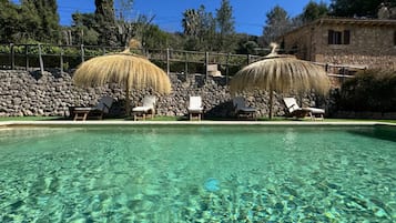 Ein klares Schwimmbecken mit Liegestühlen und Strohdächern, umgeben von Steinmauern und Bäumen.