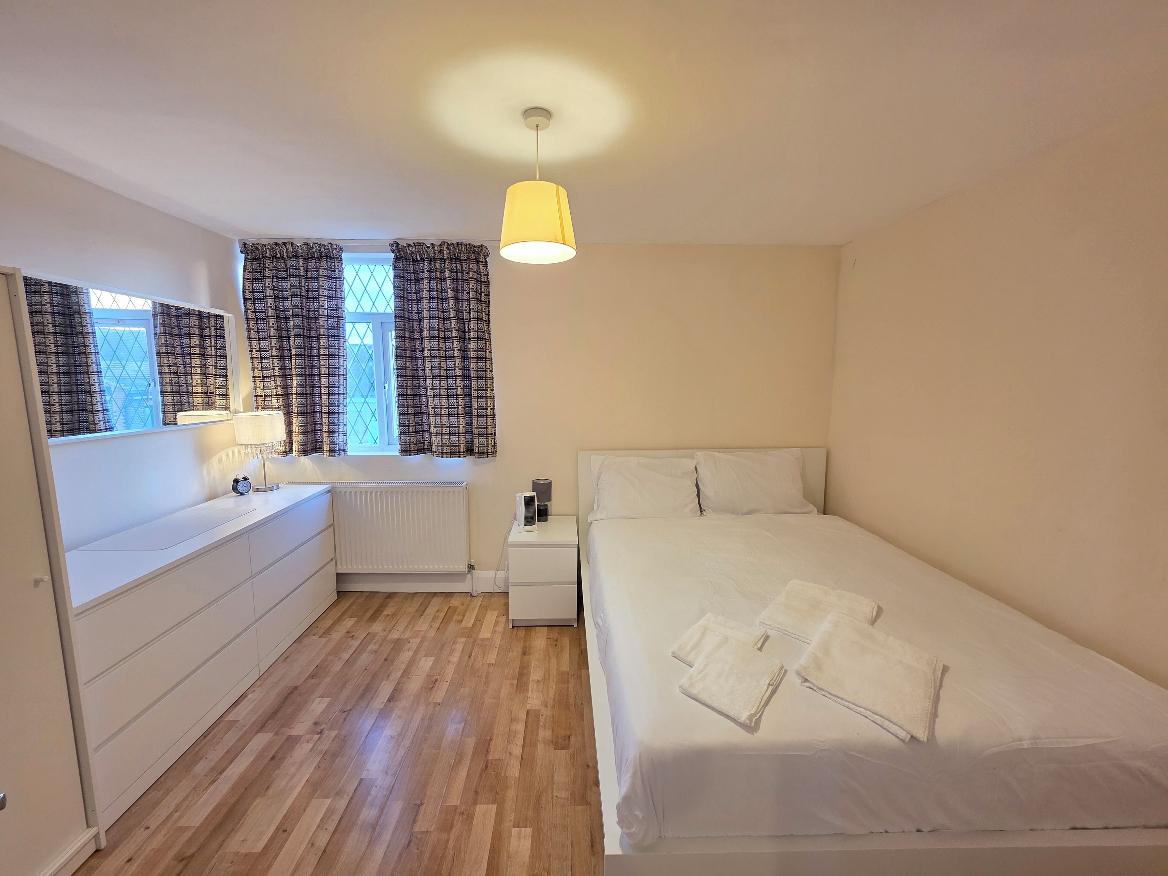 A bedroom with a bed, a bedside table, a lamp, a window with curtains, a radiator, and a wooden floor.