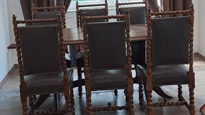 En matplats med ett träbord och stolar, stora fönster med gardiner och utsikt över grönområden utanför.