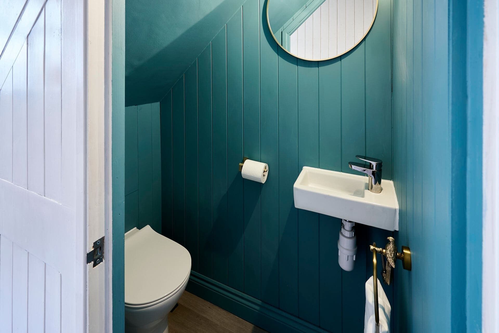 Un baño con paredes verde azulado, un inodoro blanco, un lavabo blanco con grifo cromado y un espejo redondo.