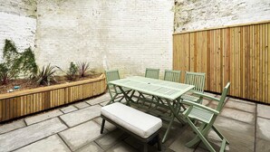 Een terras met een houten tafel en stoelen, een bankje en een plantenbak met planten.