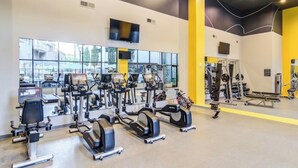 Een sportschool met diverse trainingsapparaten, een aan de muur bevestigde televisie en grote ramen.