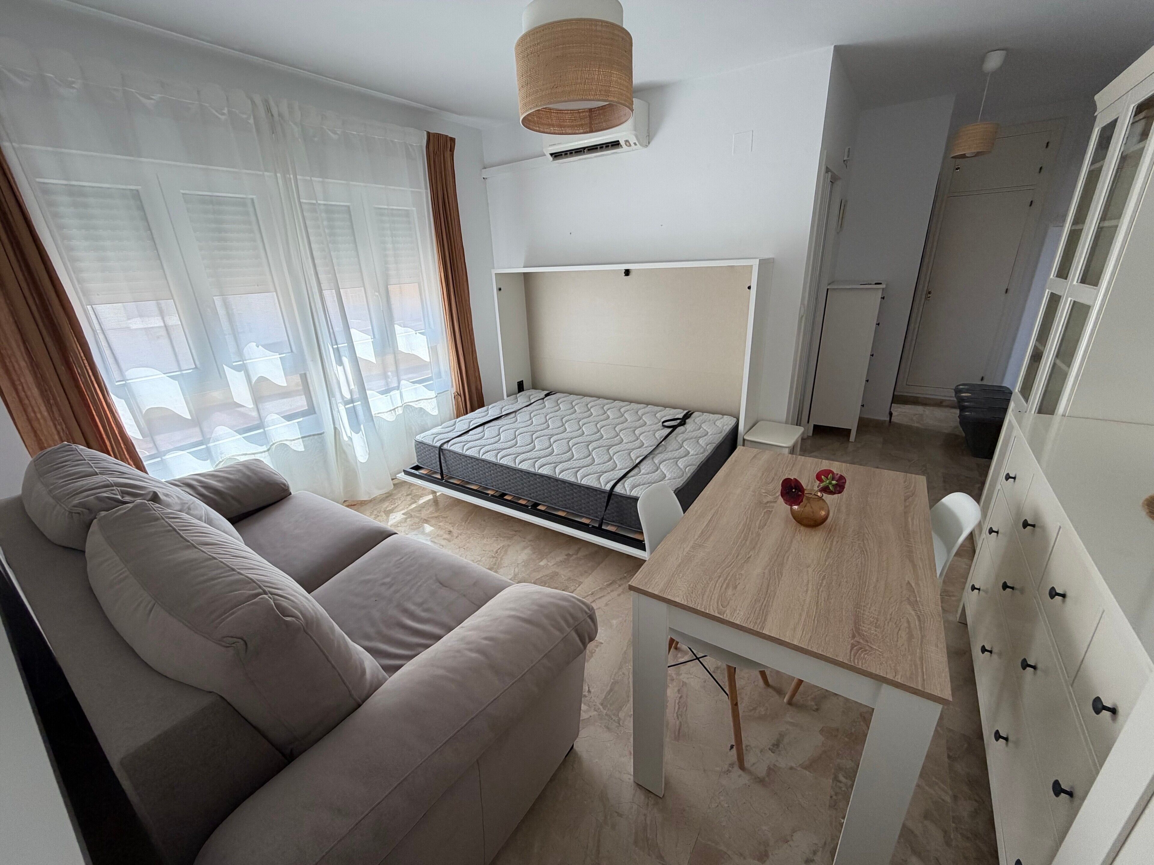 A compact room with a sofa, a small table, a bed with a mattress, a ceiling fan, and a window with curtains.