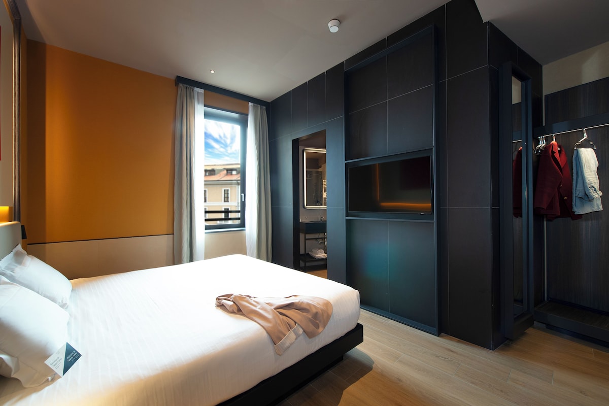 A modern hotel room with a large bed, built-in wardrobe, and a view through the window.
