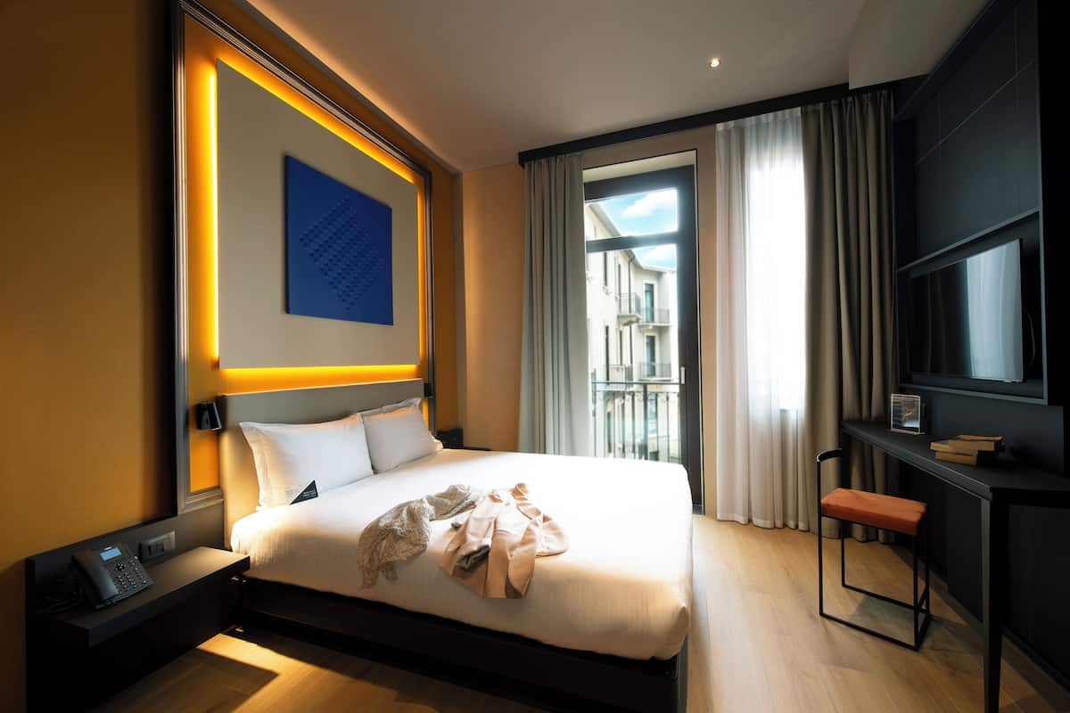 A modern hotel room with a large bed, a desk with a chair, a telephone, and a view of the outside.