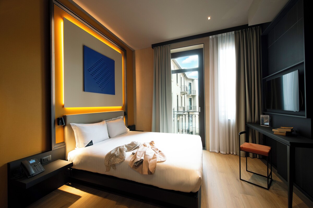 Une chambre d’hôtel moderne, dotée d’un grand lit, d’un bureau avec une chaise, d’un téléphone et offrant une vue sur l’extérieur.