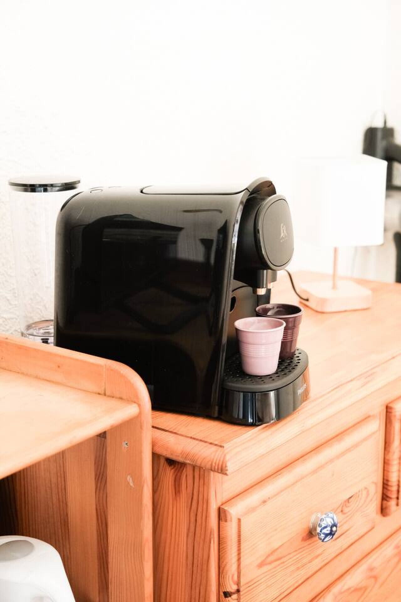 Una cafetera sobre una encimera de madera, con un porta tazas y una lámpara al fondo.