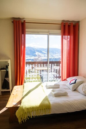 Une chambre à coucher avec un lit, un banc en bois et une vue sur des montagnes enneigées, visible à travers une grande fenêtre.