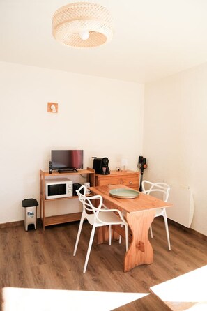 Un petit coin repas avec une table en bois et des chaises blanches, un four à micro-ondes, une machine à café et un téléviseur posé sur une étagère en bois.