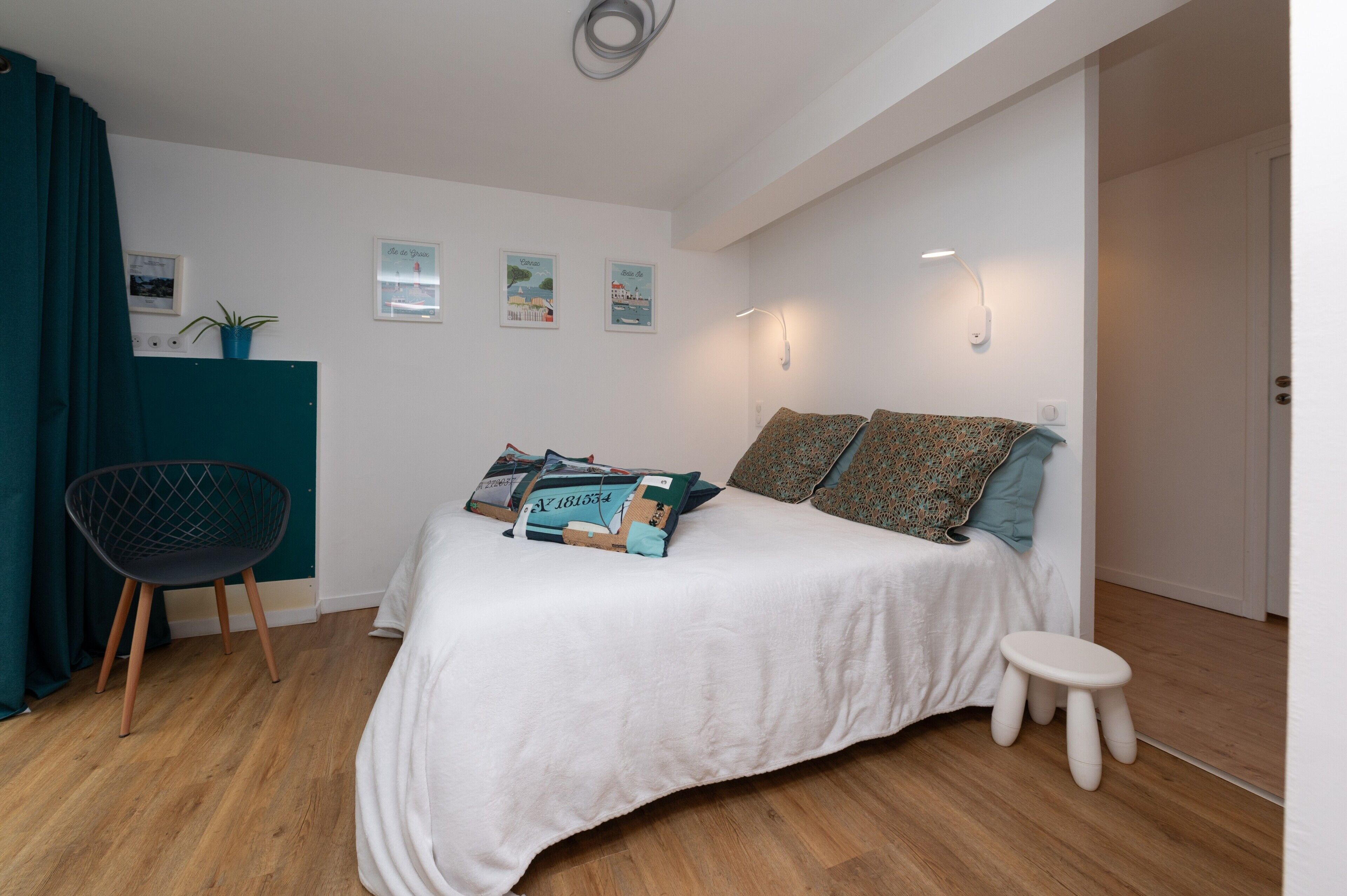 Quarto com cama, uma cadeira, um banquinho pequeno e luminárias na parede.