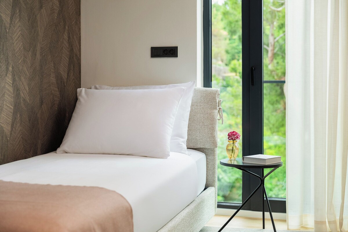 A bedroom with a bed, a small table with a vase and book, and a view of greenery through a window.