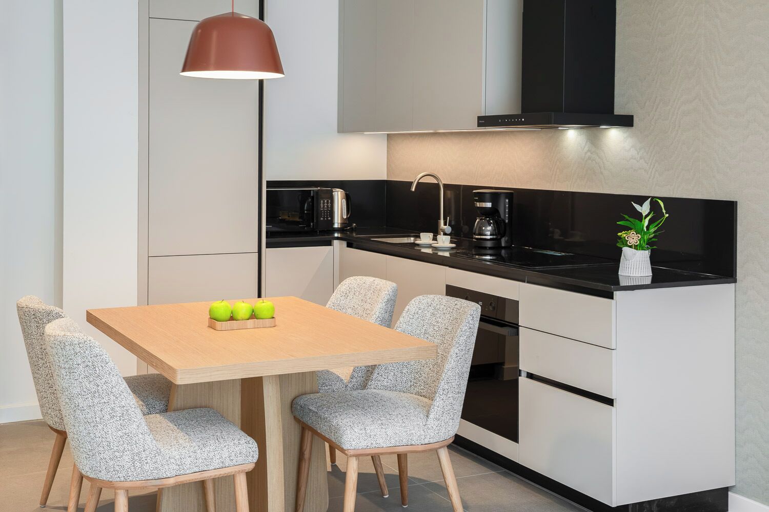 A compact kitchen with a dining area, featuring a wooden table, speckled chairs, a pendant light, and modern appliances.