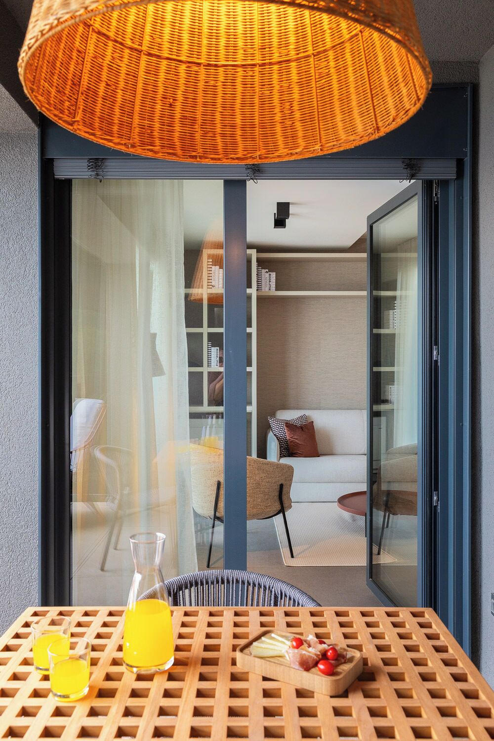 Un interior moderno con una puerta de cristal que da a una sala de estar, una lámpara de mimbre y una mesa con bebidas y aperitivos.