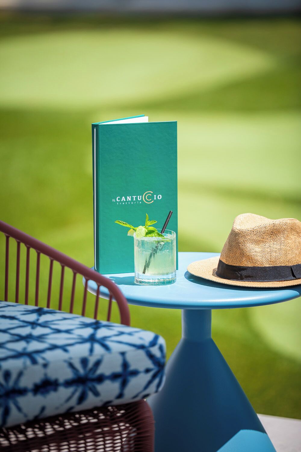 Ein Tisch mit einem Buch mit dem Titel 'CantuCio' und einem Strohhut, vor einem unscharfen grünen Hintergrund.
