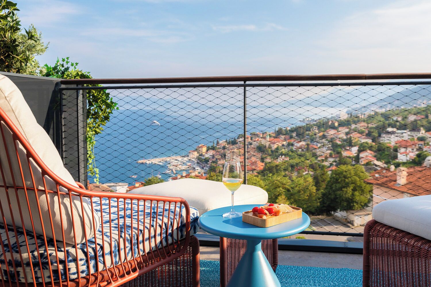 Ein Balkon mit Korbmöbeln, ein Tisch mit einem Weinglas und Obst, und Blick auf eine Küstenstadt.