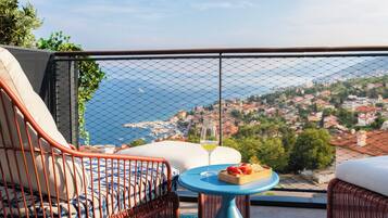 Ein Balkon mit Korbmöbeln, ein Tisch mit einem Weinglas und Obst, und Blick auf eine Küstenstadt.