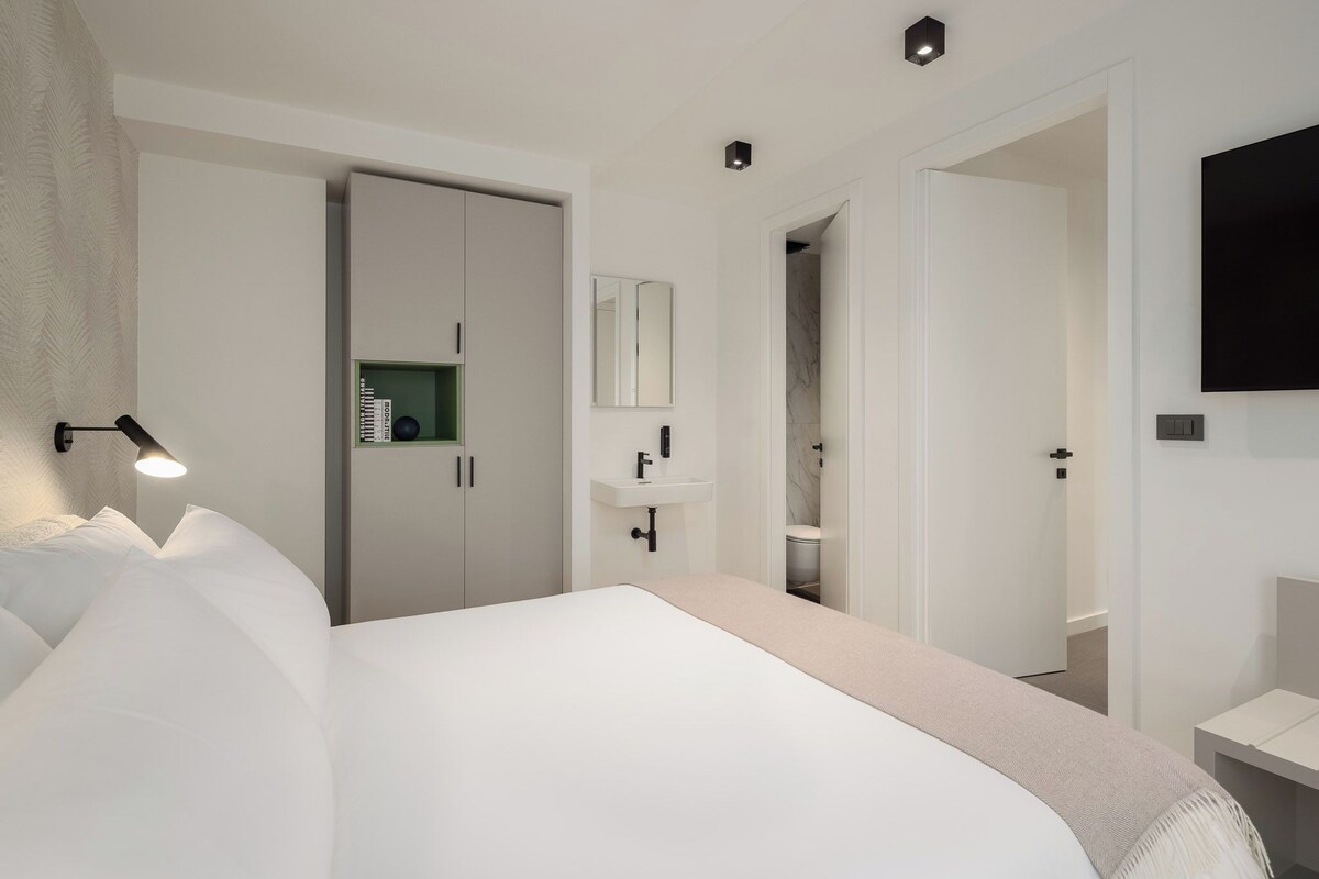 Une chambre d’hôtel moderne dotée d’un grand lit, d’une lampe de chevet, d’une armoire, d’une salle de bain avec un miroir et un lavabo, et d’un téléviseur fixé au mur.