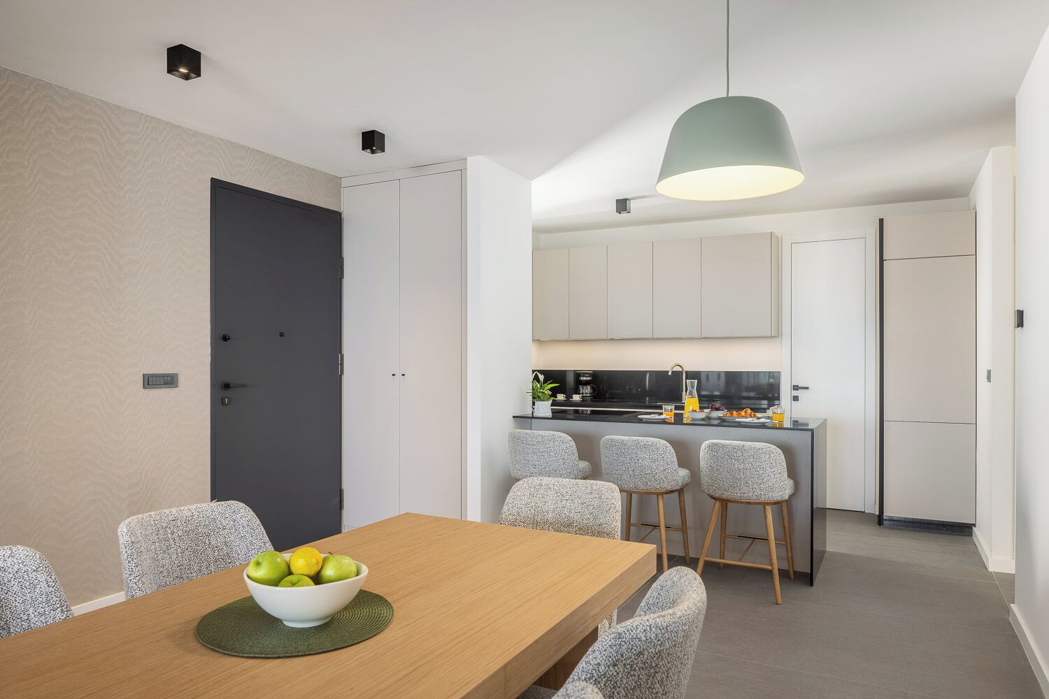 A modern kitchen with white cabinets, a central island with bar stools, and a dining table with a bowl of fruit.