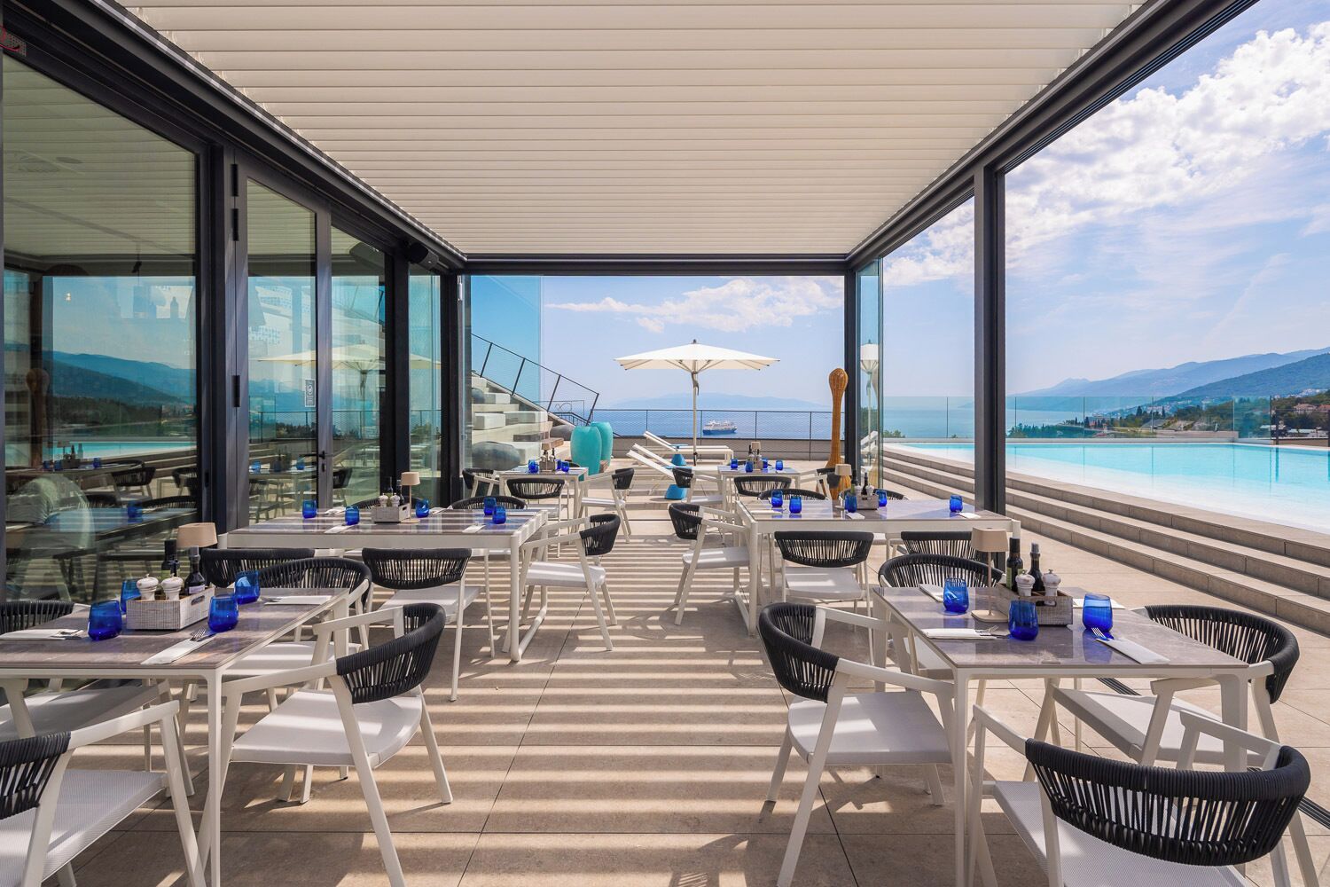 A rooftop terrace with tables and chairs, overlooking a pool and a scenic view.