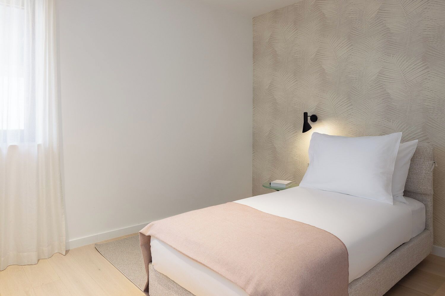 A neatly made bed with a light-colored bedspread and pillows, a bedside table with a book, and a wall-mounted lamp.