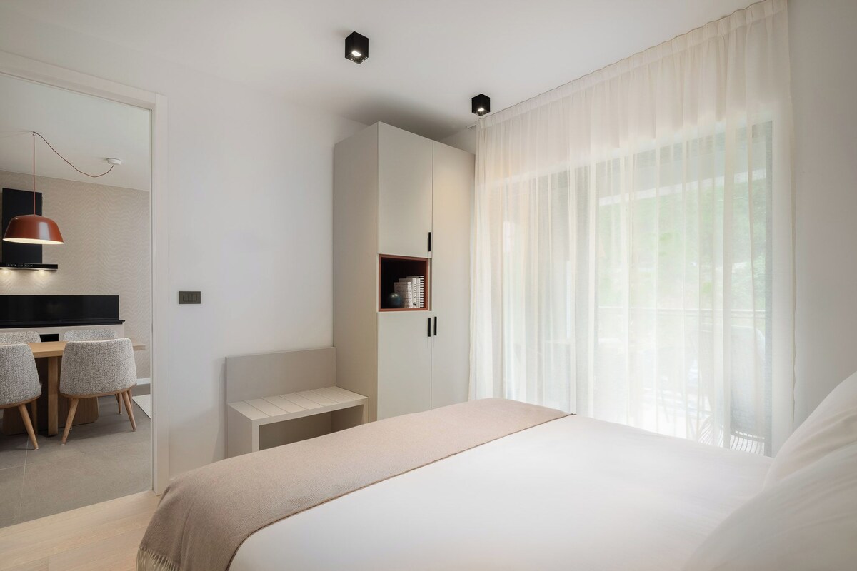 Una habitación de hotel moderna con una cama, un escritorio con silla, un armario y una ventana con cortinas translúcidas.
