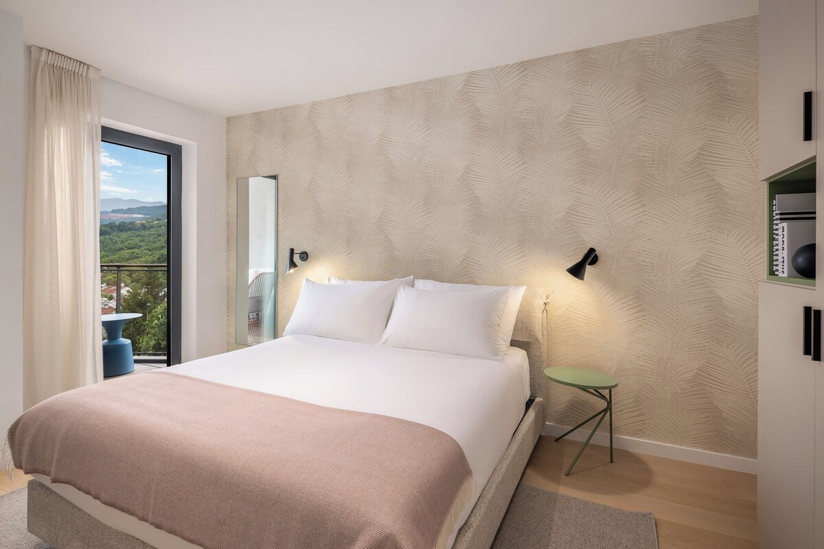Un dormitorio con cama, mesita de noche, espejo, lámpara de pared y vistas al exterior.