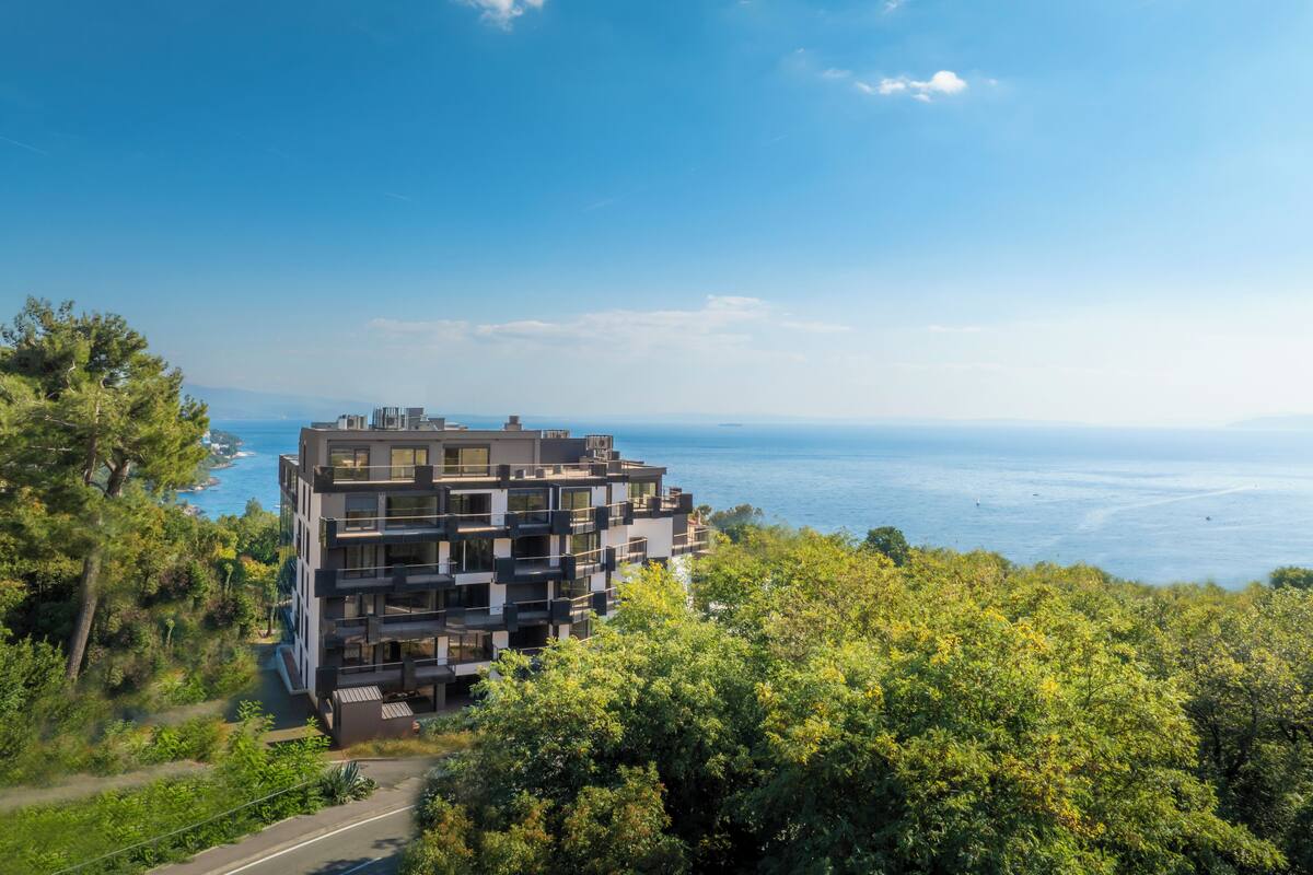 A modern multi-story building with balconies, surrounded by lush greenery and overlooking a body of water.